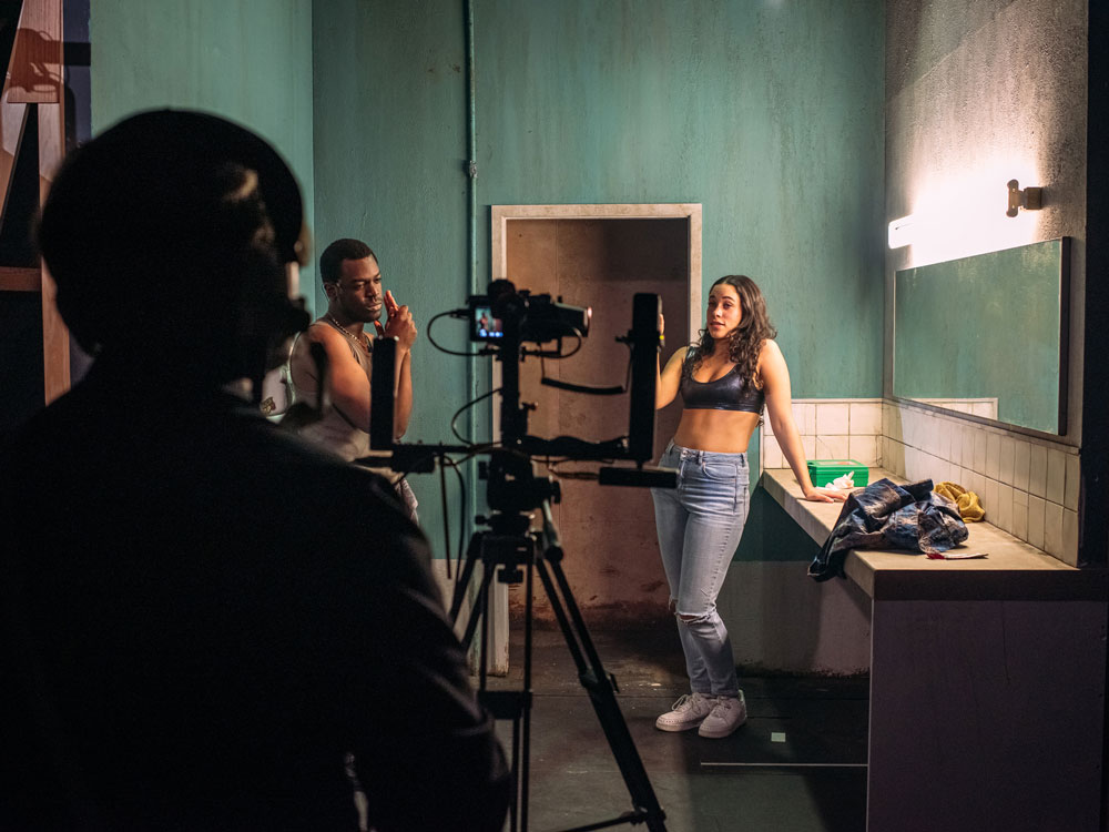 两个演员站在一个俱乐部的浴室里，而一个制作艺术系的学生在操作摄像机
